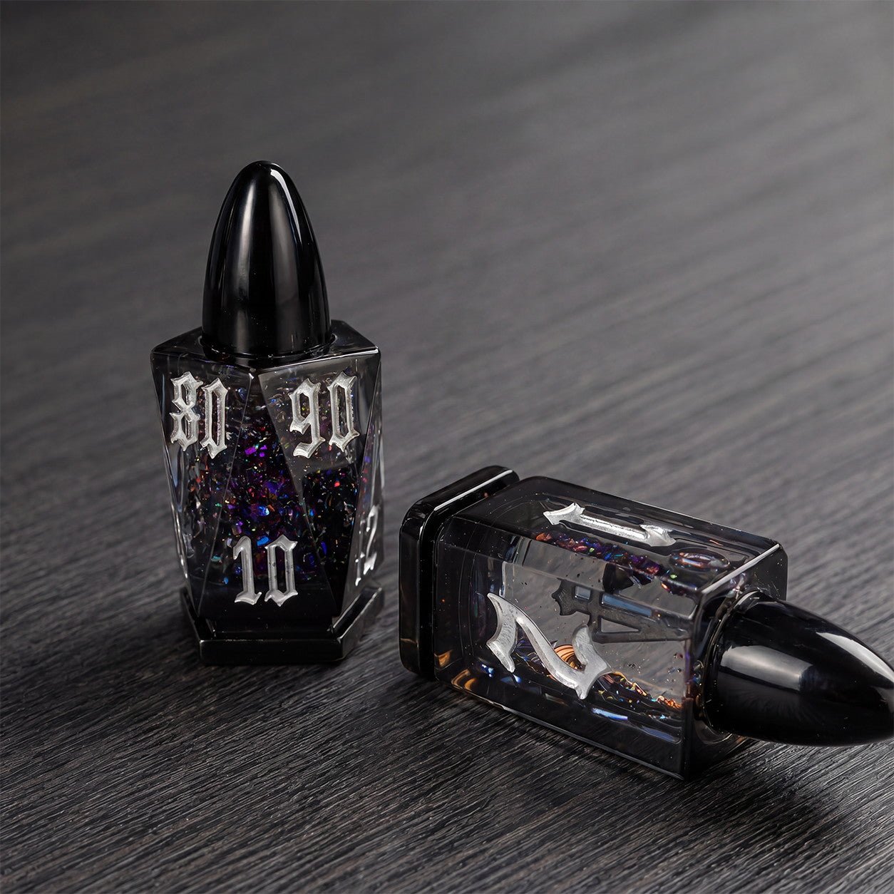 Midnight Assassin close-up of two black bullet-shaped liquid core DND dice with iridescent flakes on a dark tabletop.