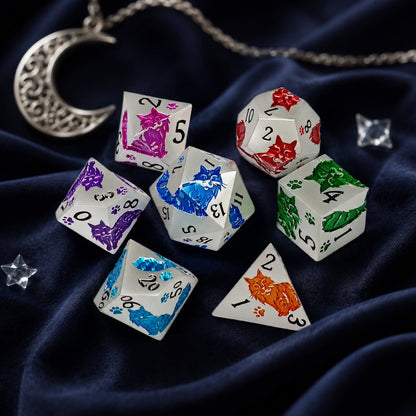 Moonwhisker 7-piece metal DND dice set with colorful cat motifs on deep blue velvet beside a moon charm.