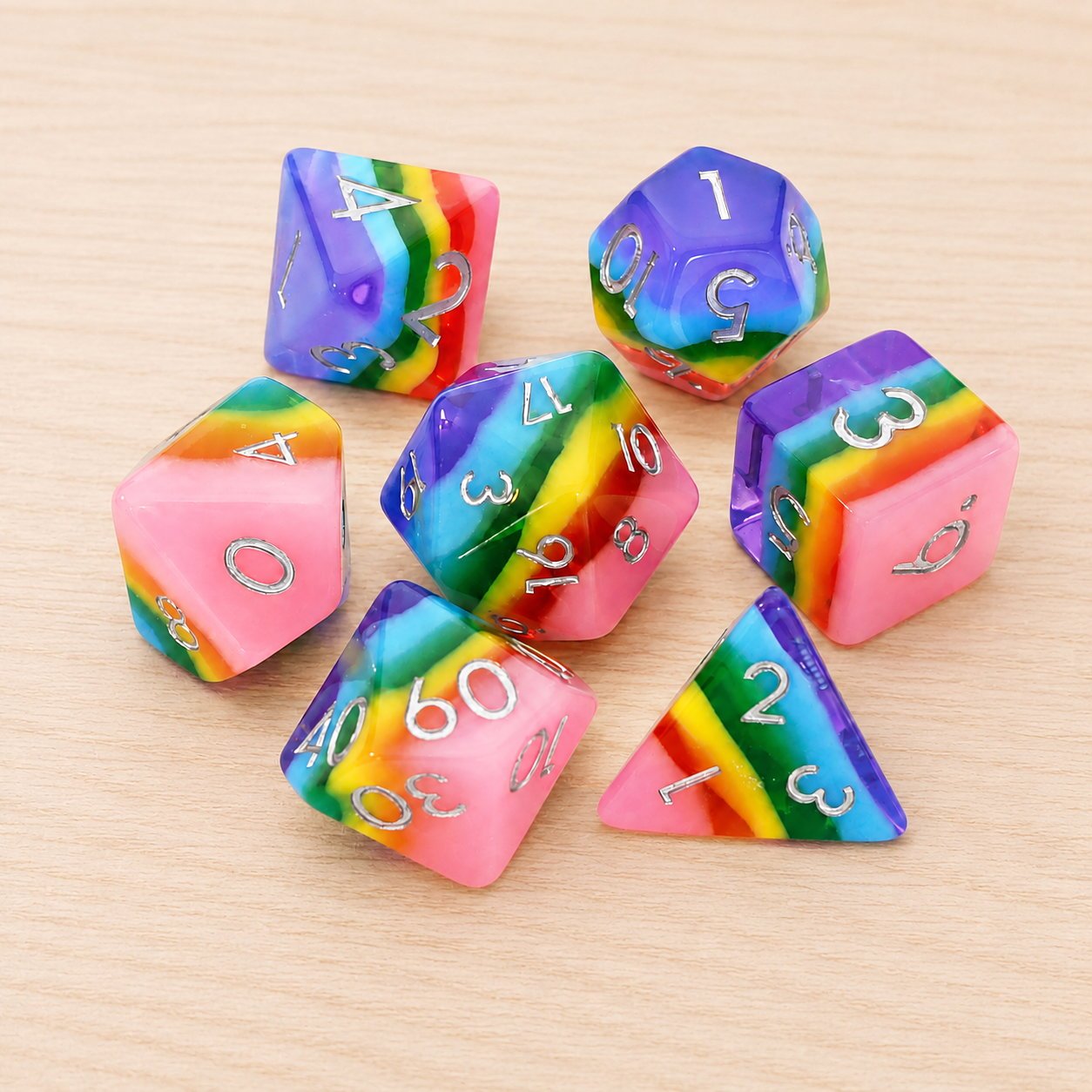 Rainbow Candy 7-piece rainbow layered resin DND dice set with silver numbers on a light wood tabletop.